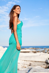 Beauty in teal dress poses confidently on rocky coast, embodying elegance and transformation.