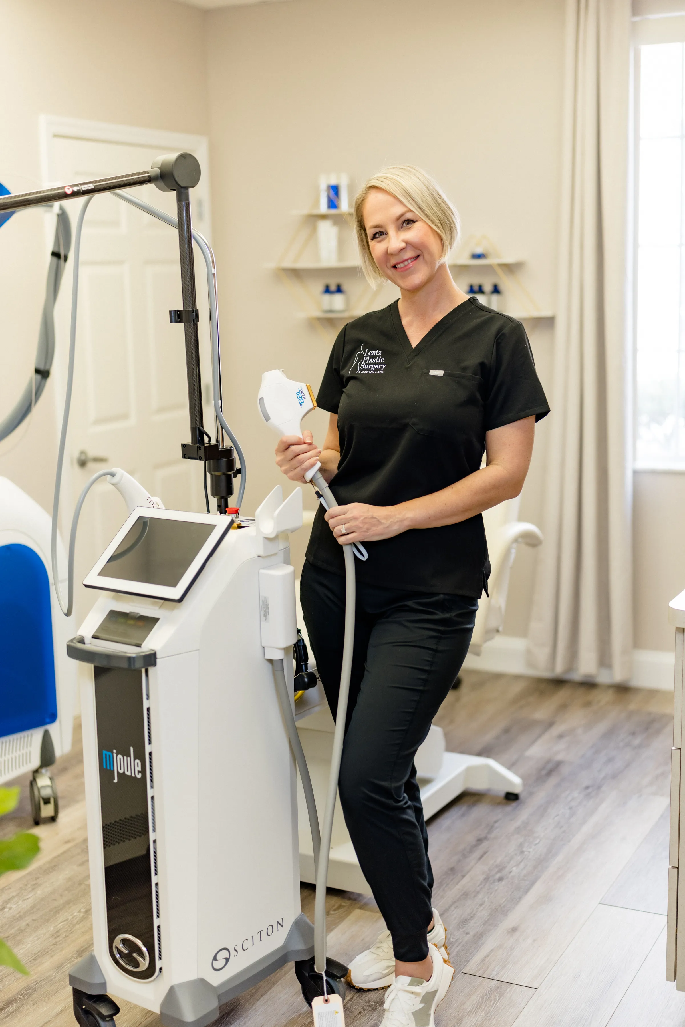Judit Professional aesthetician in black scrubs posing with a medical-grade laser treatment device at an aesthetic clinic.